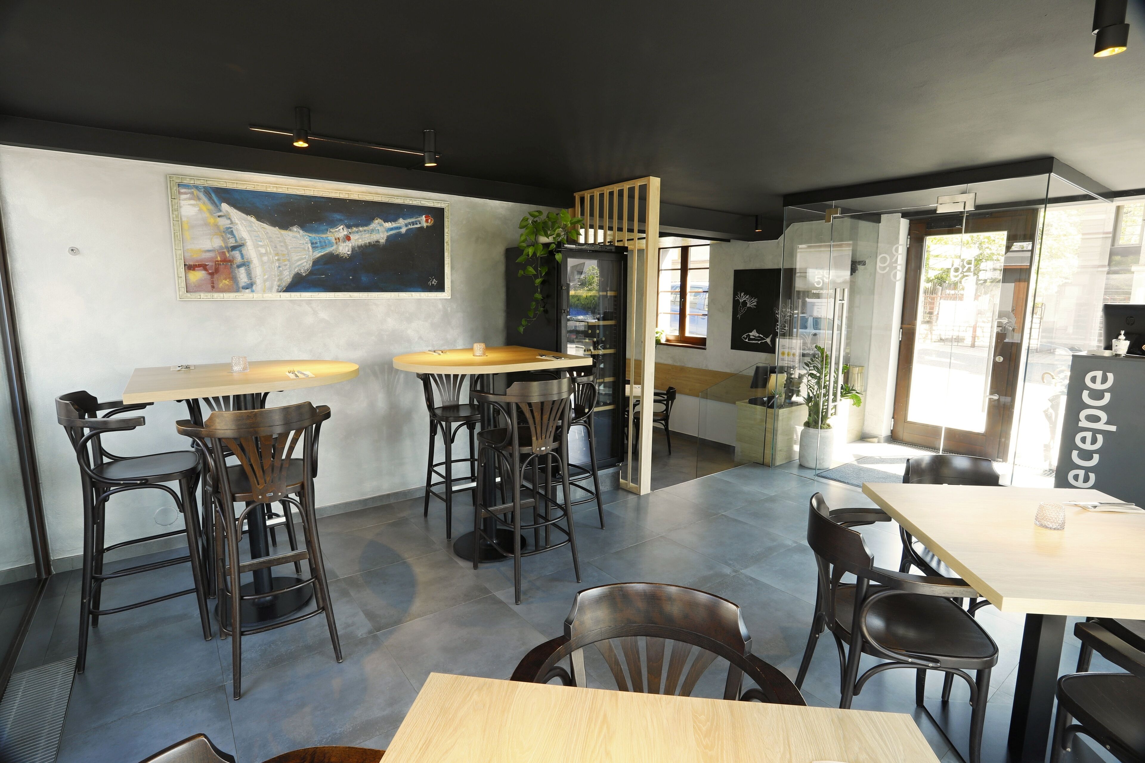 Un restaurante moderno con mesas y sillas altas, un cuadro grande en la pared y un separador de vidrio.