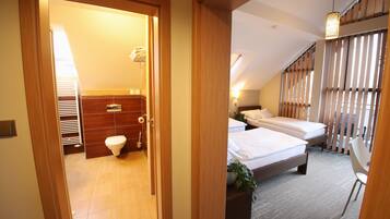 Een hotelkamer met een houten deur naar een badkamer, een bed met een plant en een raam met jaloezieën.