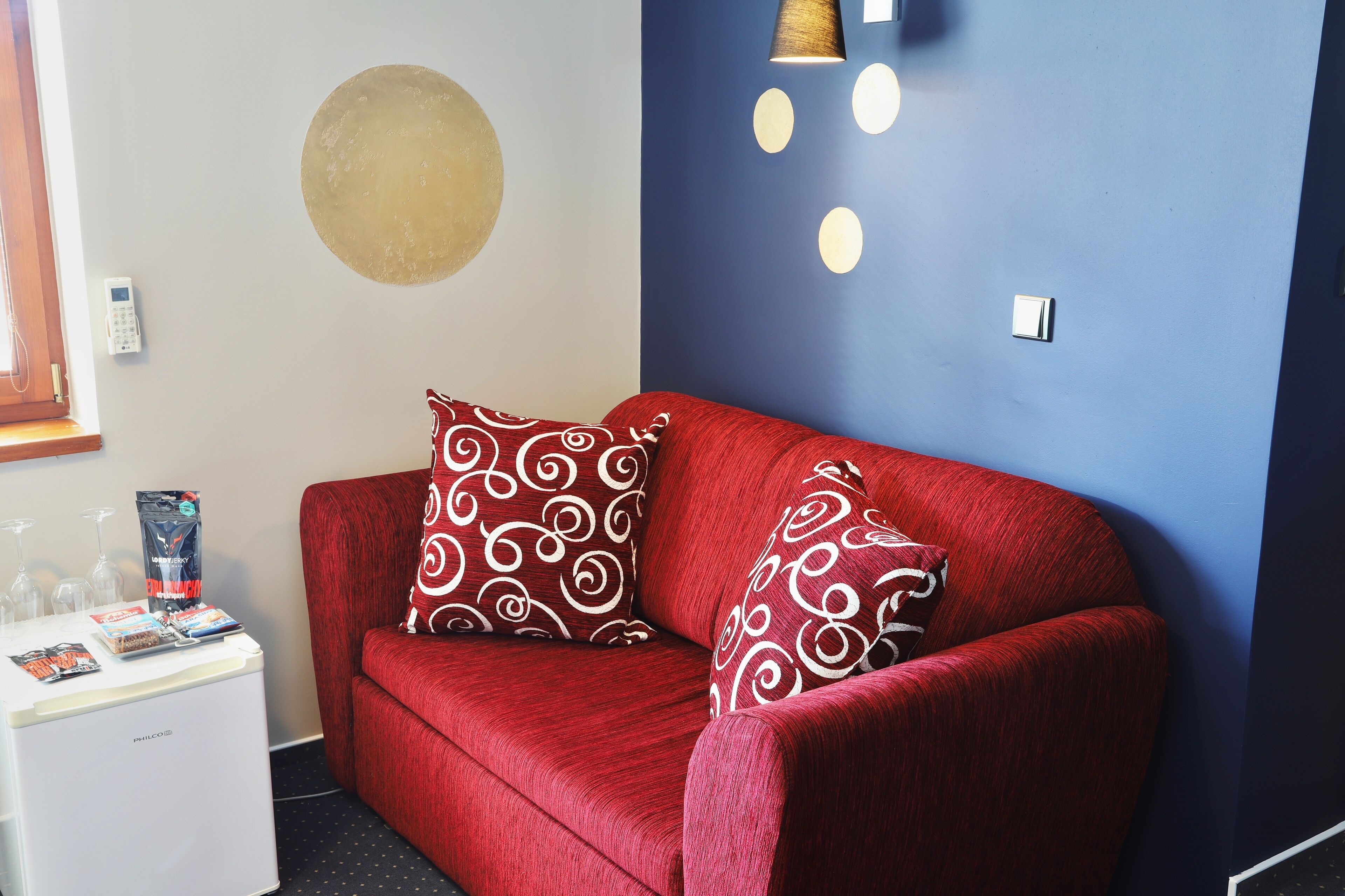 Ein rotes Sofa mit gemusterten Kissen, ein kleiner weißer Beistelltisch mit Zeitschriften, eine Wanduhr und eine runde Wanddekoration.