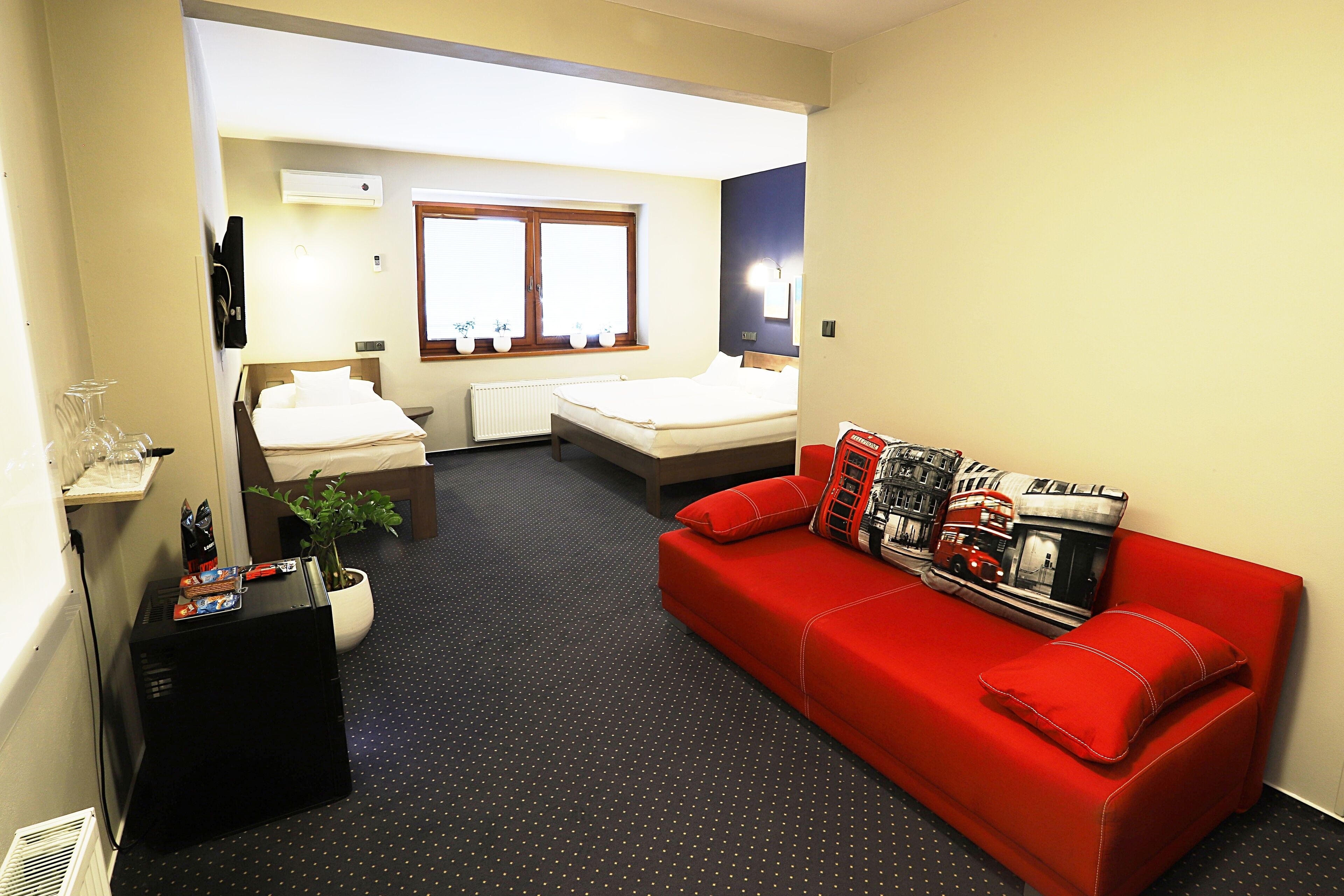 A compact hotel room with a bed, a red sofa, a small table with a plant, and a radiator.