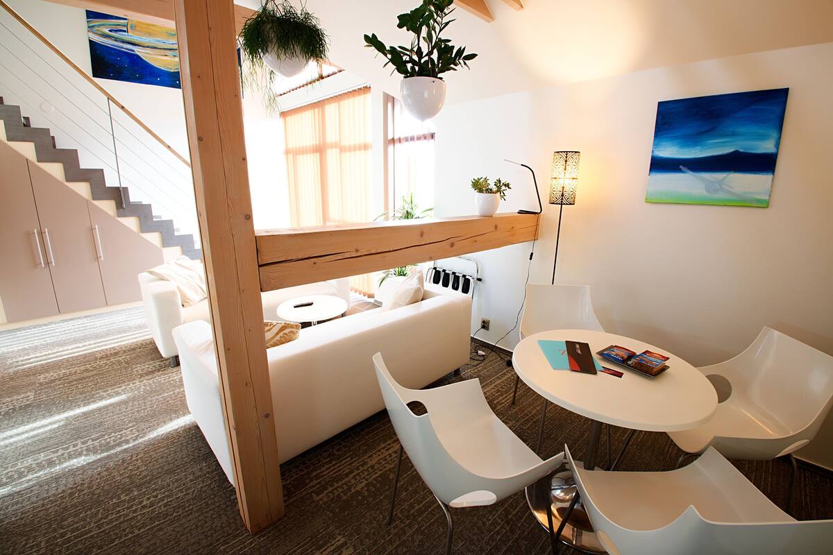 Una camera d'albergo moderna con soffitto in legno, un divano bianco, sedie bianche, un tavolino rotondo e un quadro appeso al muro.