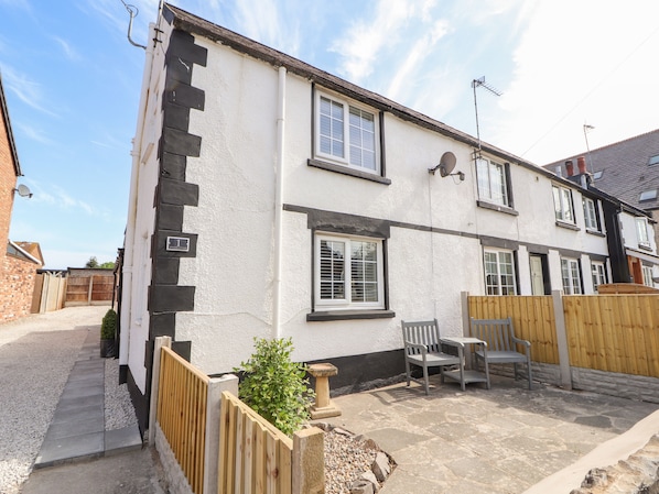 A two-story house with a white exterior and black trim. There are two windows on the ground floor and one on the upper floor. A small patio area with two chairs and a table is visible. - Afallon Cottage (Rhyl)