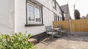 Un bâtiment blanc avec une fenêtre, une clôture en bois et une terrasse en pierre avec deux chaises.