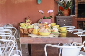 Une table de petit-déjeuner dressée avec du pain, des viennoiseries et des boissons, entourée de chaises blanches, dans une pièce dont un mur est en briques et qui est décorée de plantes en pot.