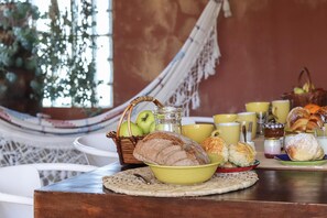 Desayuno con pan, bollería y una cesta de fruta sobre una mesa.