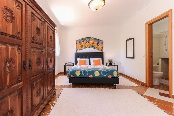 A bedroom with a large wooden wardrobe, a bed with a colorful polka dot bedspread, and a headboard with a decorative top.