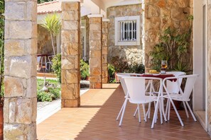 Un patio con paredes de piedra, sillas y una mesa blancos, rodeado de plantas y un suelo de baldosas.
