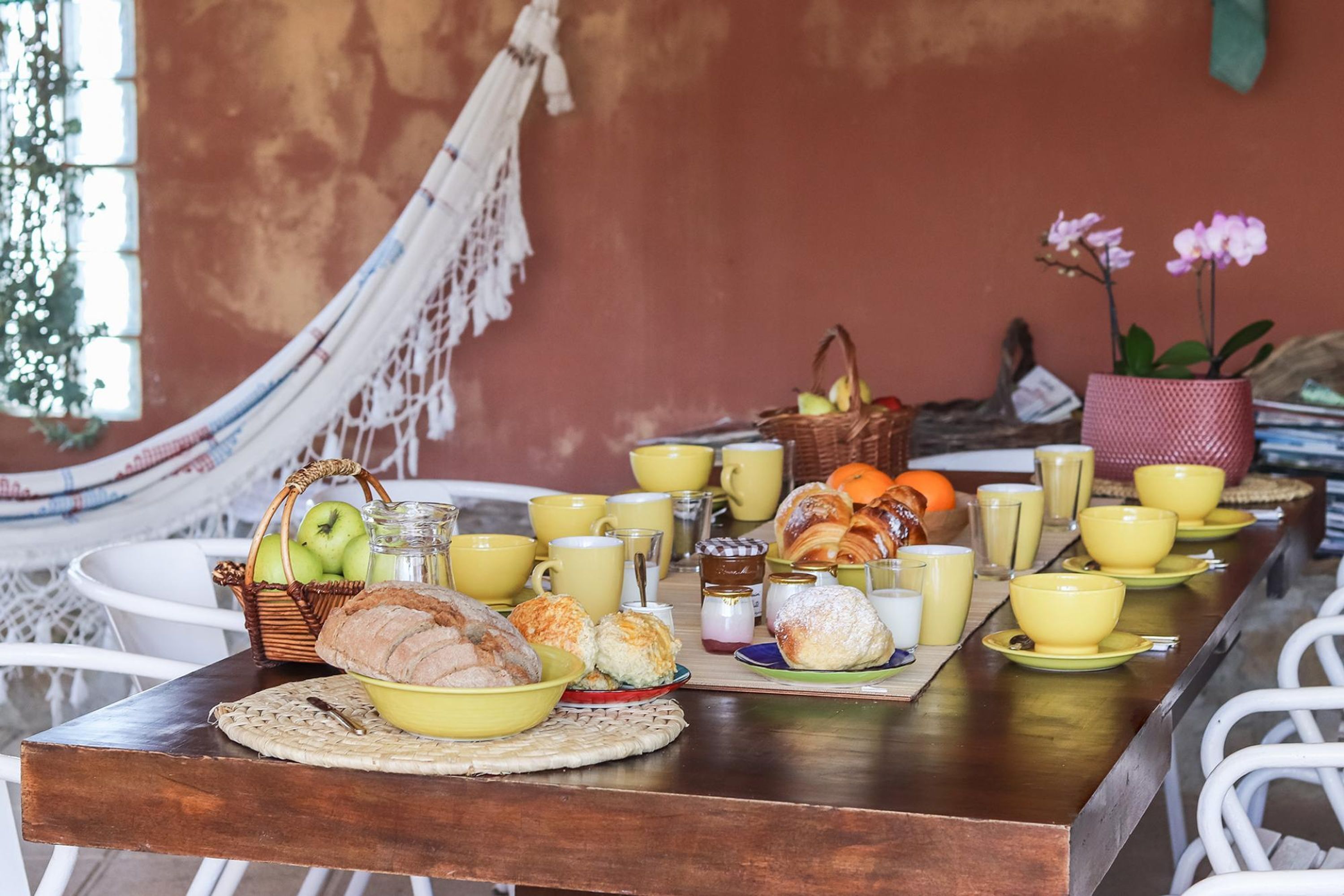 Una mesa de desayuno con diversos alimentos, tazas amarillas y una hamaca al fondo.