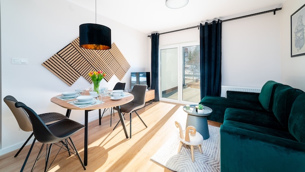 Ein modernes Wohnzimmer mit einem grünen Sofa, einem Essbereich mit einem Holztisch und Stühlen, einer schwarzen Pendelleuchte und einem großen Fenster mit Vorhängen.