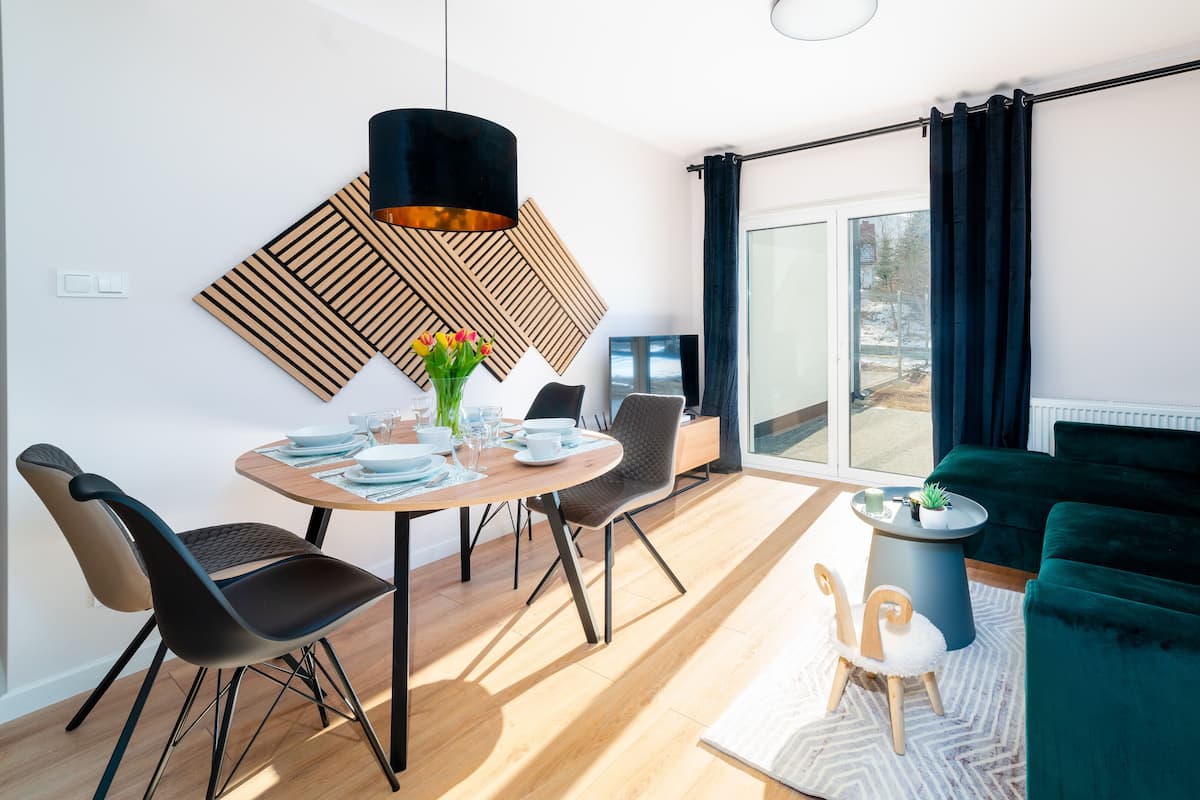 Un salon moderne avec un canapé vert, un coin repas avec une table et des chaises en bois, un luminaire suspendu noir et une grande fenêtre avec des rideaux.
