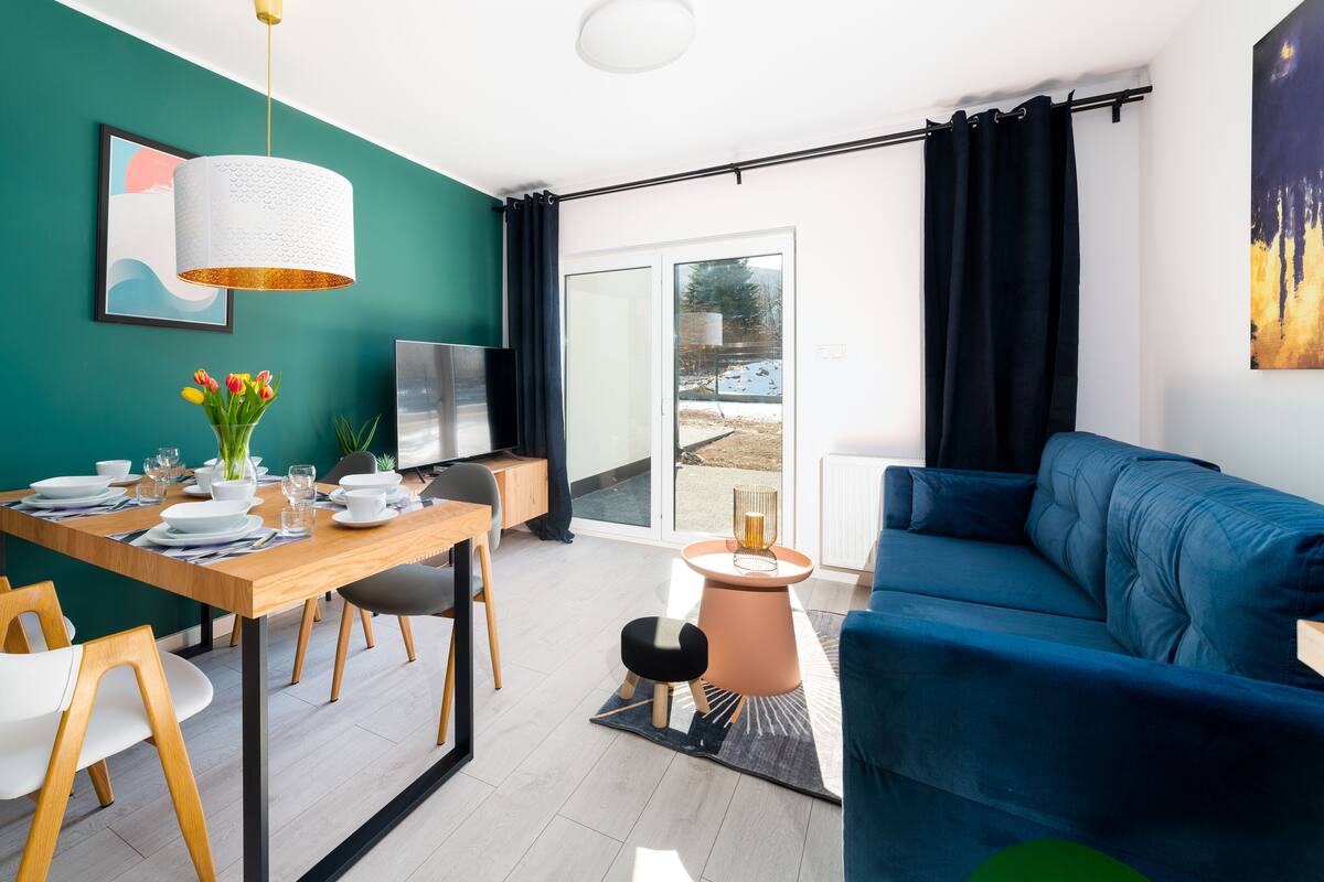 Un salon moderne avec un canapé bleu, une table à manger en bois mise avec des assiettes et des verres, un téléviseur et un vase de tulipes.