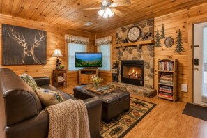 A cozy log cabin interior with a fireplace, television, leather furniture, and a bookshelf.