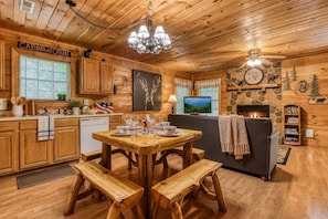 A cabin interior with a wooden dining table, benches, a fireplace, a television, and a kitchen area.