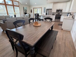 A dining area with a wooden table and chairs, a kitchen in the background, and large windows.