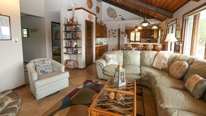 A cozy living room with a sofa, armchair, and a coffee table. There is a bookshelf, a dining area, and a kitchen visible in the background. - Tara - Enjoy easy beach access from this Traditional 3 BR Beach Cottage in Southern Shores (Southern Shores)