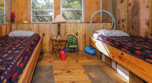 Une petite chambre avec lits superposés, située sous la maison et protégée par des moustiquaires, comprenant deux lits, une petite table, des chaises et une fenêtre donnant sur les arbres.