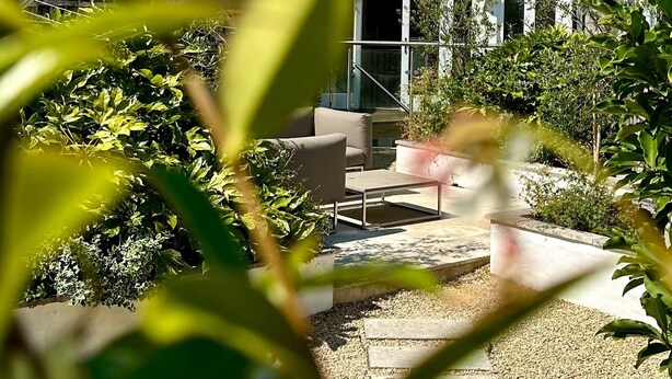 Una casa a due piani con facciata bianca, un camino in vista e un piccolo giardino con patio e posti a sedere.