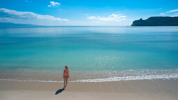 Eine Frau steht an einem Strand mit Blick auf das Meer und ein fernes Gebirge.