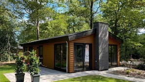 A modern log cabin with large glass doors and windows, surrounded by a well-maintained lawn and trees.