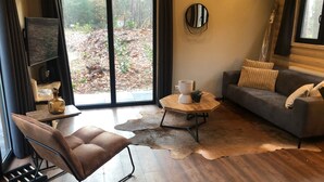 A modern living room with a leather chair, a grey sofa, a round coffee table, and a large window with a view of a wooded area.
