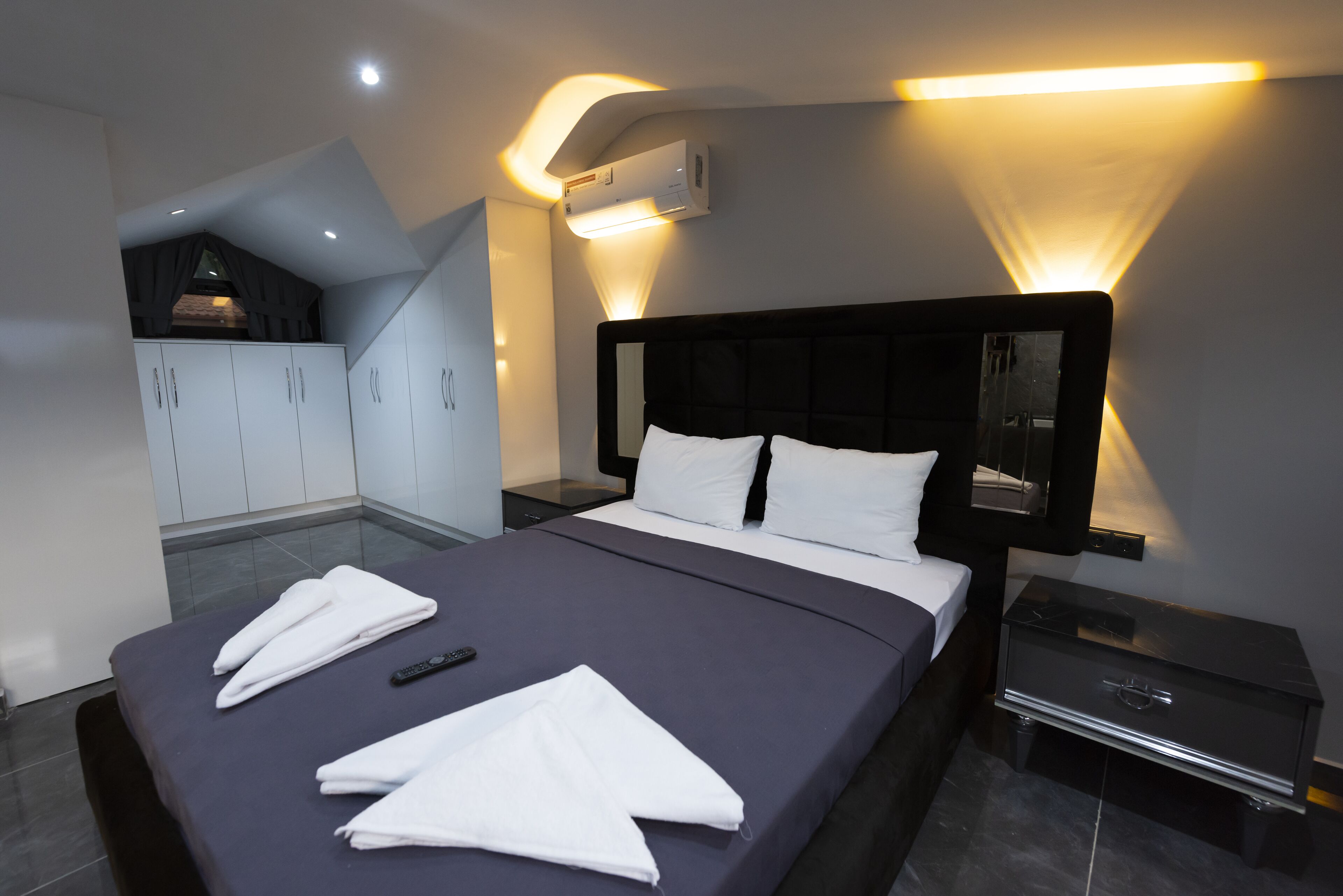 A modern hotel room with a large bed, a nightstand, a wardrobe, and an air conditioning unit.