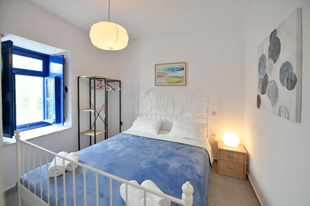 A bedroom with a bed, a bedside table with a lamp, a window with blue curtains, a wall with a painting, and a shelf unit.