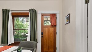 A room with a wooden door, a window with green curtains, a small table with a plant, and a chair.