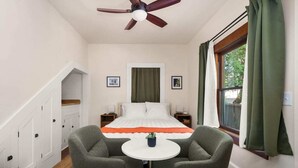 A bedroom with a bed, two chairs, a small table, a ceiling fan, a window with curtains, and a staircase.