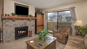 A living room with a stone fireplace, a flat-screen TV, a comfortable sofa, and a large window with a view of buildings. - Prime Avon Location | Walk to Gondola & Dining | River Trail Access & Views (Avon)