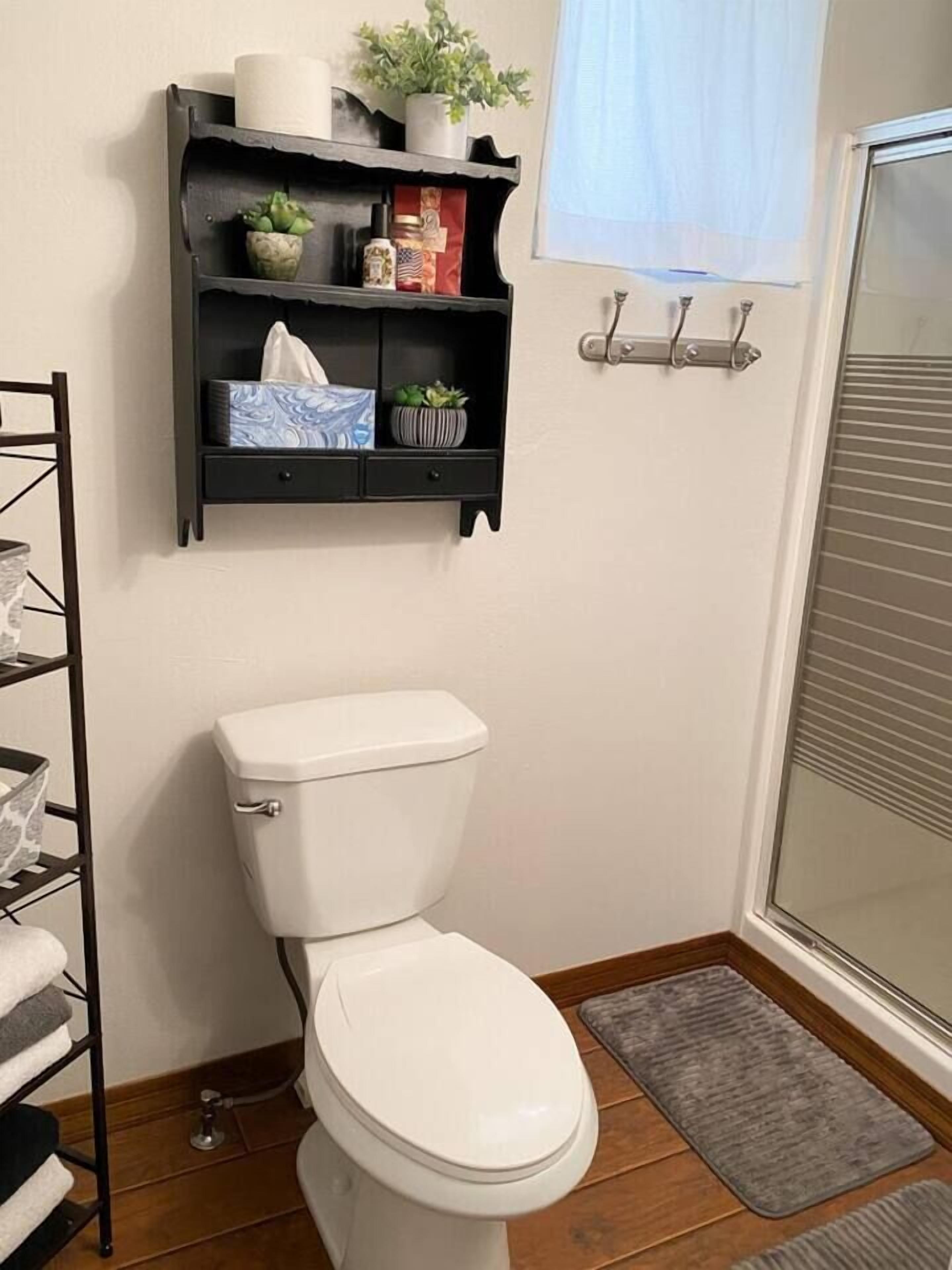 A bathroom with a white toilet, a shelf with toiletries, a small plant, and a window with blinds.