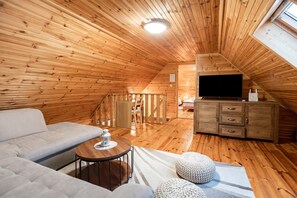 A cozy attic bedroom with a wooden sloped ceiling, a sofa, a small round table, a TV, and a dresser.