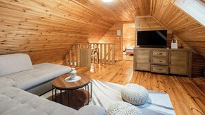 A cozy attic room with wooden walls and a sloped ceiling. It features a sofa, a small round table, and a TV mounted on the wall. - Chalet Markoci With Hot Tub (Rakovica)