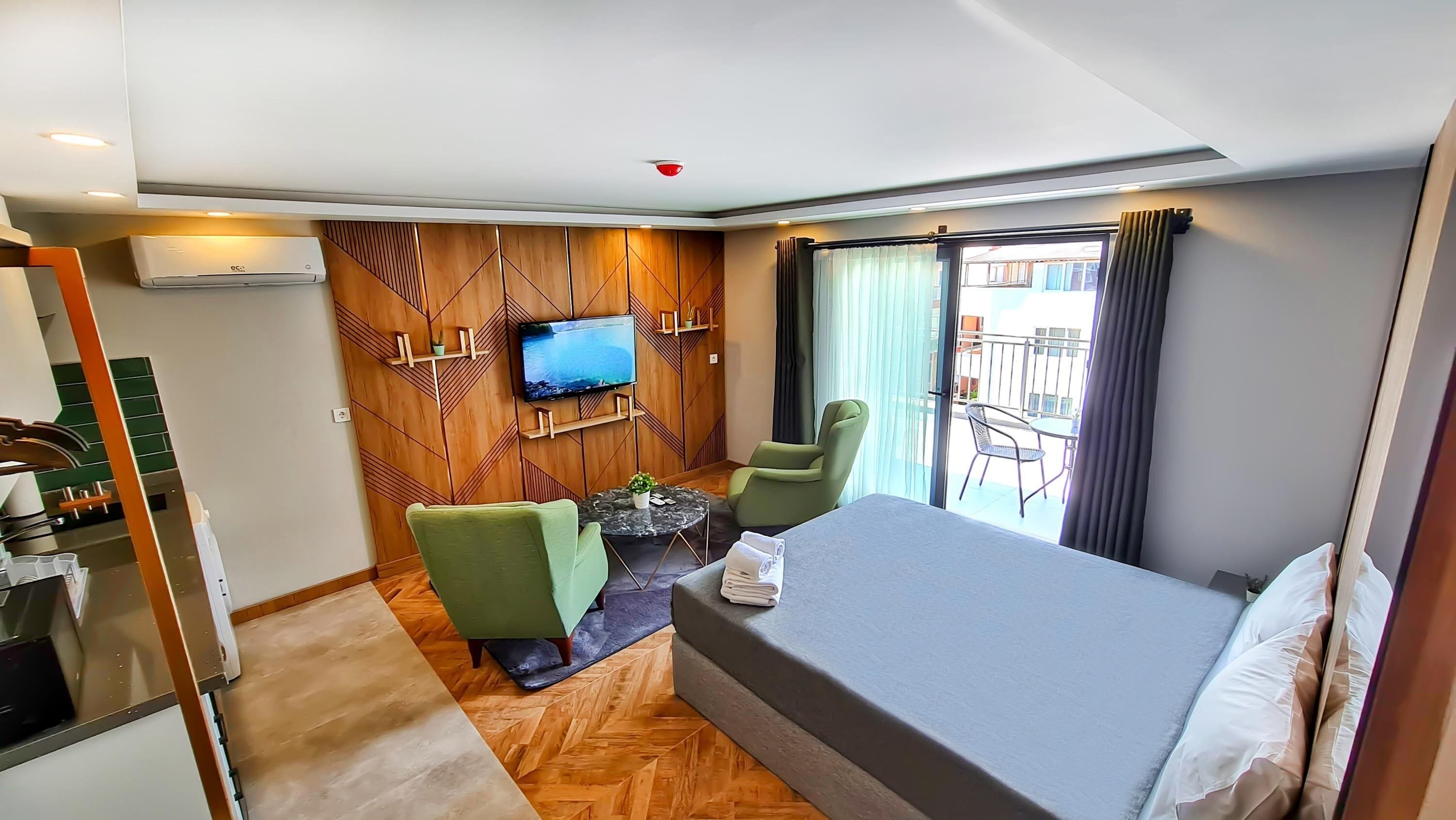 A hotel room with a bed, a TV mounted on a wooden panel wall, a small table with a plant, two green chairs, and a balcony with a table and chairs.