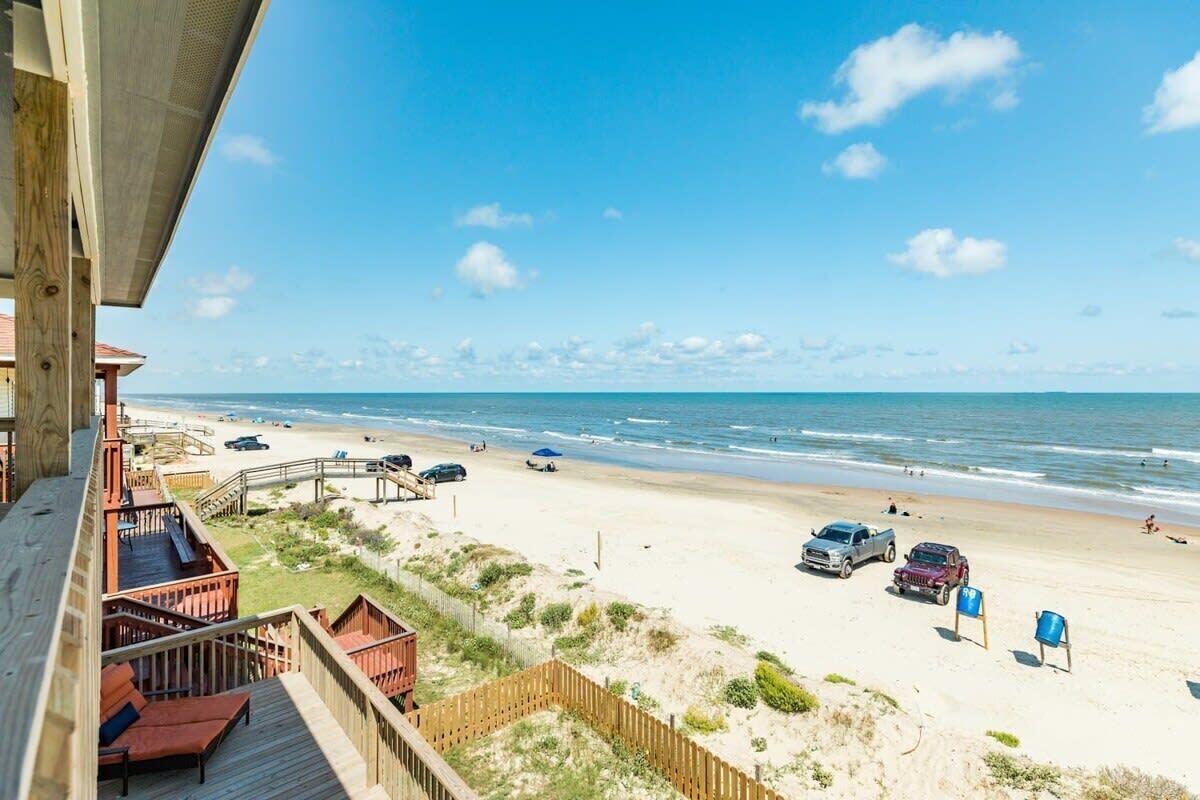 Une vue sur la plage depuis un balcon doté d’une terrasse en bois, avec une plage de sable et un ciel bleu dégagé.
