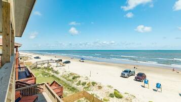 Une vue sur la plage depuis un balcon doté d’une terrasse en bois, avec une plage de sable et un ciel bleu dégagé.