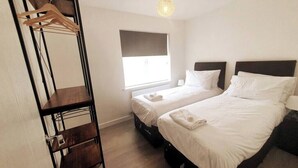 A hotel room with two beds, a wooden wardrobe, a window with blinds, and a hanging light fixture.