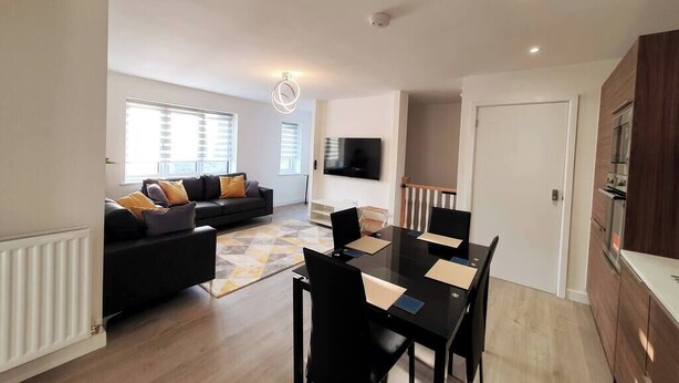 A modern living room with a black sofa, a dining table with chairs, a flat-screen TV mounted on the wall, and a kitchen area with wooden cabinets.