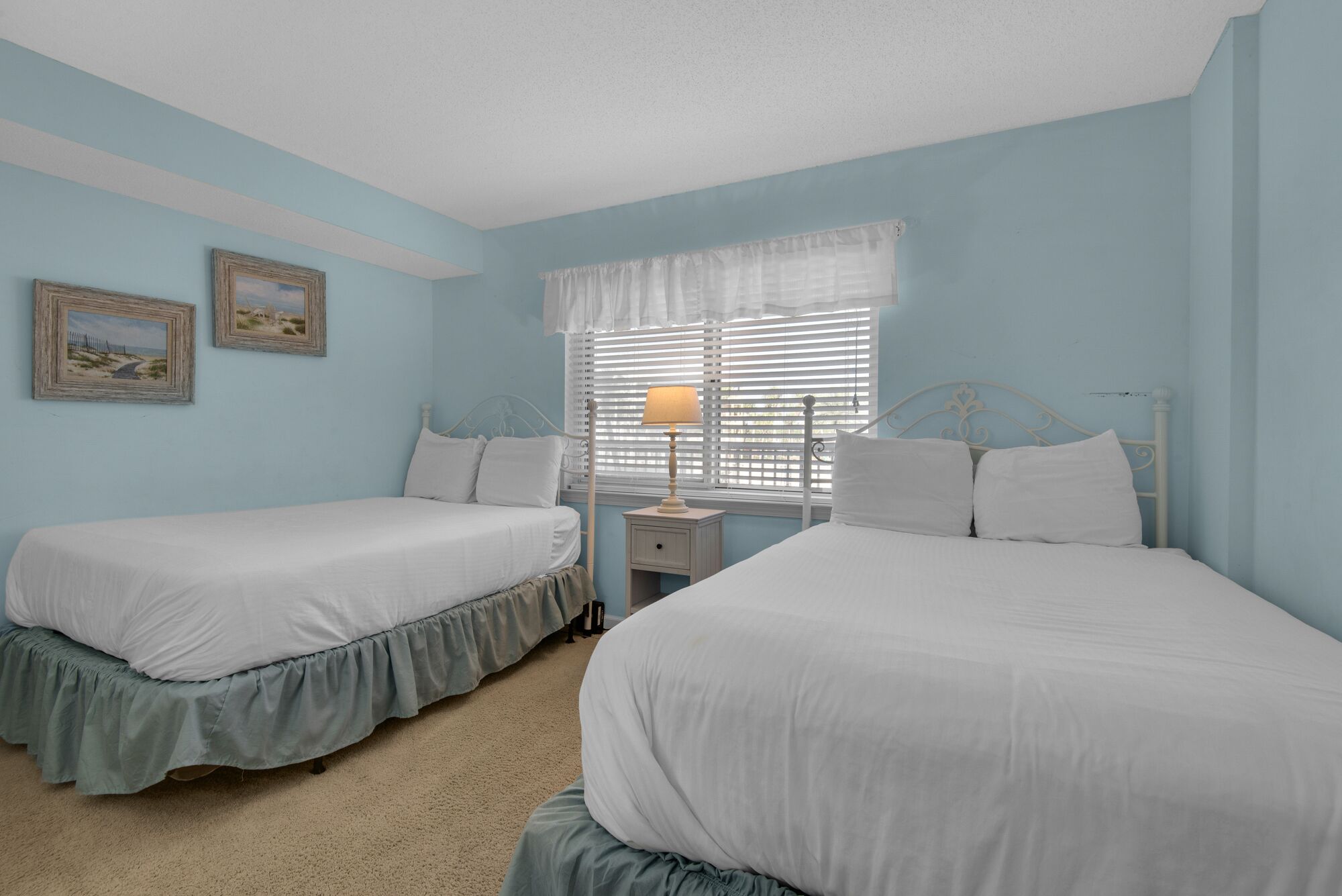 A bedroom with two single beds, a nightstand with a lamp, and a window with blinds.