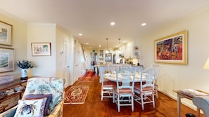 A dining area with a round table and white chairs, a kitchen in the background, and a hallway leading to a door.
