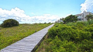 A wooden boardwalk leading to a house with a balcony, surrounded by greenery and a clear sky.
