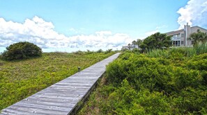 A wooden boardwalk leading to a house with a balcony, surrounded by greenery and a clear sky.