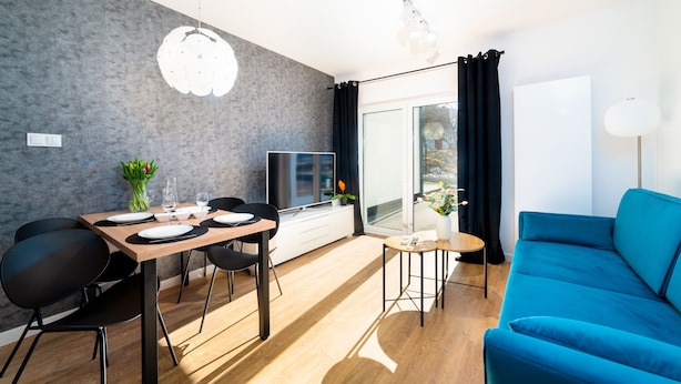 Un salon moderne avec un canapé bleu, une table à manger dressée avec des assiettes et des verres, un téléviseur et une grande fenêtre avec des rideaux.