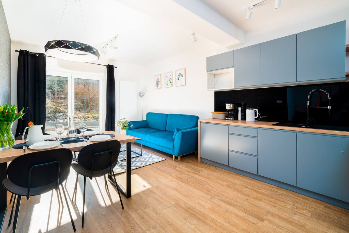 A modern kitchen with grey cabinets, a black sink, and a coffee machine. There is a blue sofa and a dining table with chairs.