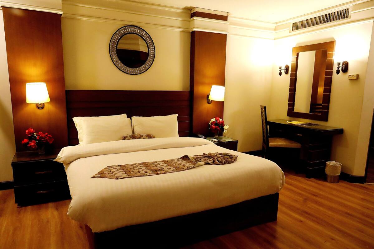 Habitació d'hotel amb un llit gran, dues taules de nit amb làmpades, un taulell, un mirall i un terra de fusta.