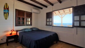 A bedroom with a bed, a nightstand, a lamp, a surfboard decoration, and a window with a view. - Sea-view villa with pool jacuzzi – sleeps 8 (Les Anses-d´Arlet)