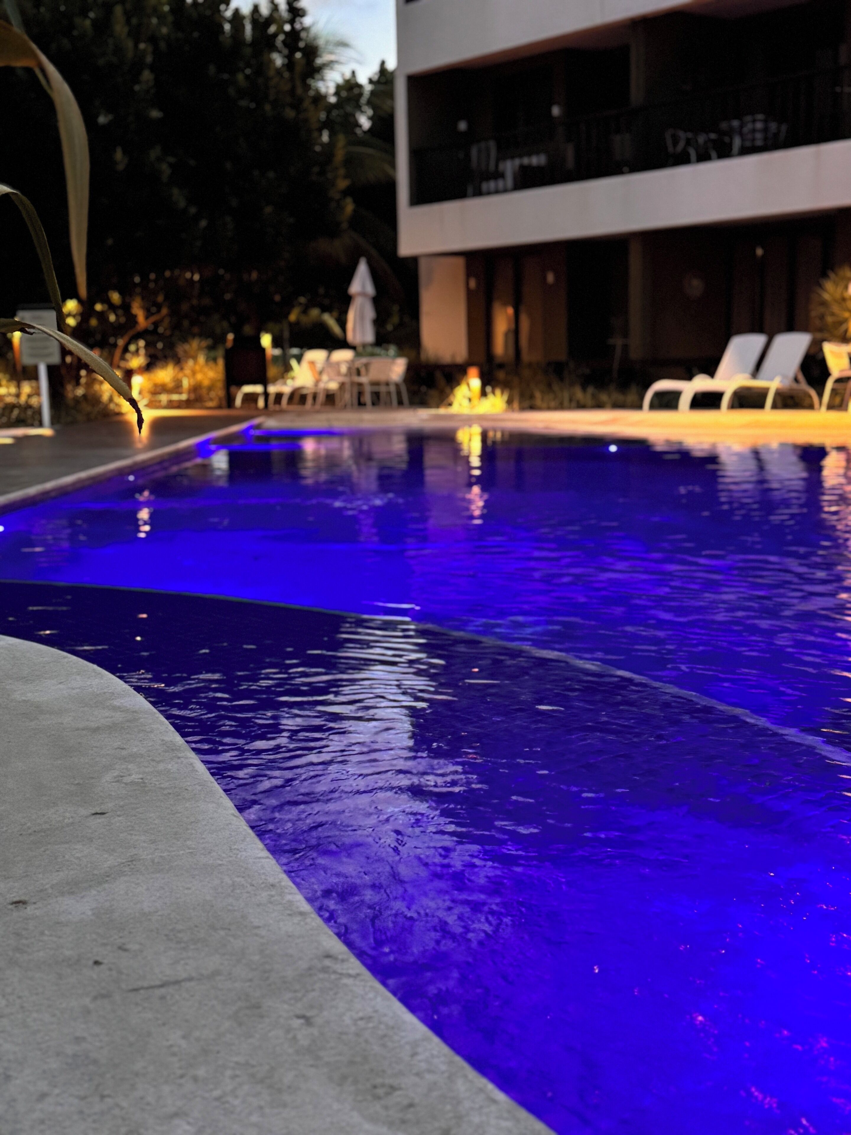 Una piscina con iluminación azul, sillas de descanso y un edificio moderno al fondo.