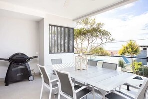 A balcony with a dining table and chairs, a grill, and a view of trees and buildings.