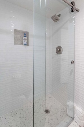 Une douche fermée par des parois en verre, dotée d’un pommeau de douche à main, d’un robinet mural et d’une petite étagère sur laquelle se trouve une bouteille.