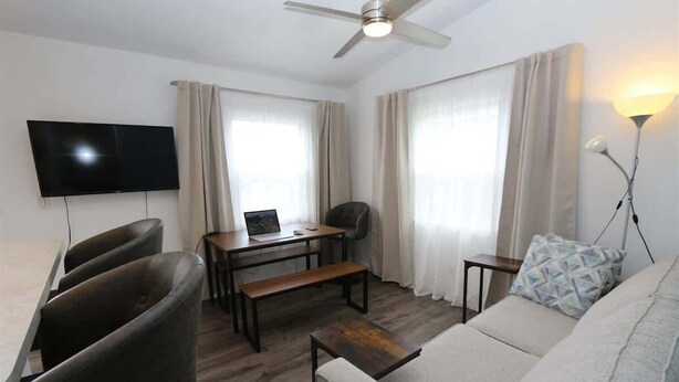 A modern living room with a flat-screen TV, a ceiling fan, a sofa, a coffee table, a desk with a laptop, and a bench.