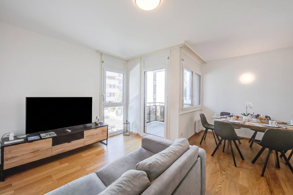 A modern living room with a flat-screen TV, a sofa, a dining table with chairs, and large windows.